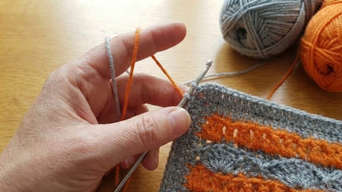 mãos de uma pessoa fazendo crochê sobre uma mesa de madeira clara. A mão esquerda segura uma peça de crochê com padrões ondulados em cinza e laranja, enquanto tensiona dois fios de lã sobre o dedo indicador. Uma agulha de metal está inserida no ponto atual. Ao fundo, há dois novelos de lã, um cinza e um laranja vibrante.
