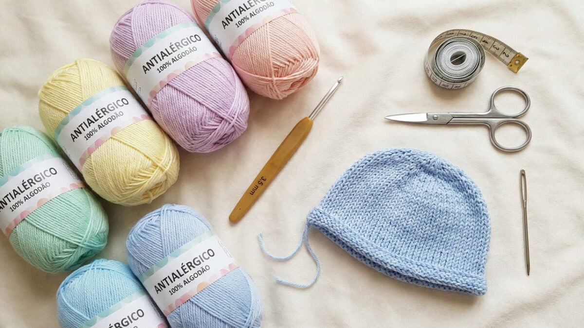 A imagem exibe diversos novelos de linha em tons pastéis (verde menta, amarelo, lavanda, rosa e azul) com rótulos onde se lê "ANTIALÉRGICO 100% ALGODÃO". No centro, há uma touca de bebê azul claro em processo de finalização, uma agulha de crochê com cabo de bambu indicando 3,5mm, uma fita métrica enrolada, uma tesoura de metal pequena e uma agulha de tapeçaria.