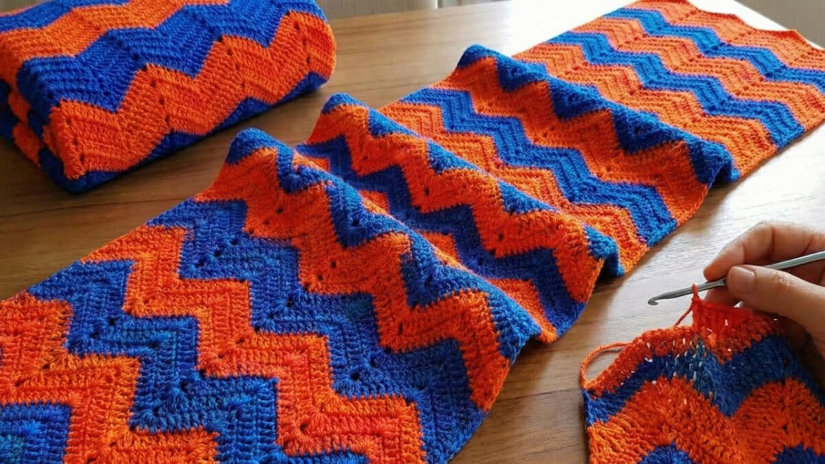 close-up de um trabalho de crochê em andamento sobre uma mesa de madeira clara. A peça utiliza a técnica "Ponto Chama", apresentando um padrão de ziguezague ondulado e fluido. As cores se alternam em listras horizontais de azul escuro, e laranja. No canto inferior direito, uma mão segura uma agulha de crochê metálica, finalizando uma das fileiras.
