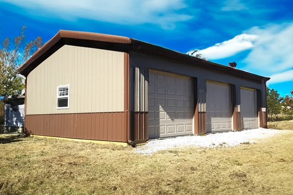 The image shows a rectangular post-frame building with a dual-tone exterior: light beige upper walls and dark brown lower siding and roof. It features three large garage doors, indicating its use as a workshop or storage facility. A small window is presen