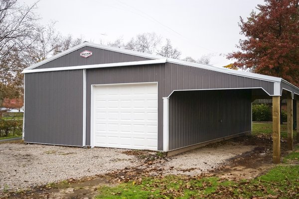 The image shows a post-frame building by Built-Mor Buildings, featuring a dark gray exterior with white trim. It includes a large white garage door, indicating its potential use as a garage or storage facility. The structure has a gable roof and an attach