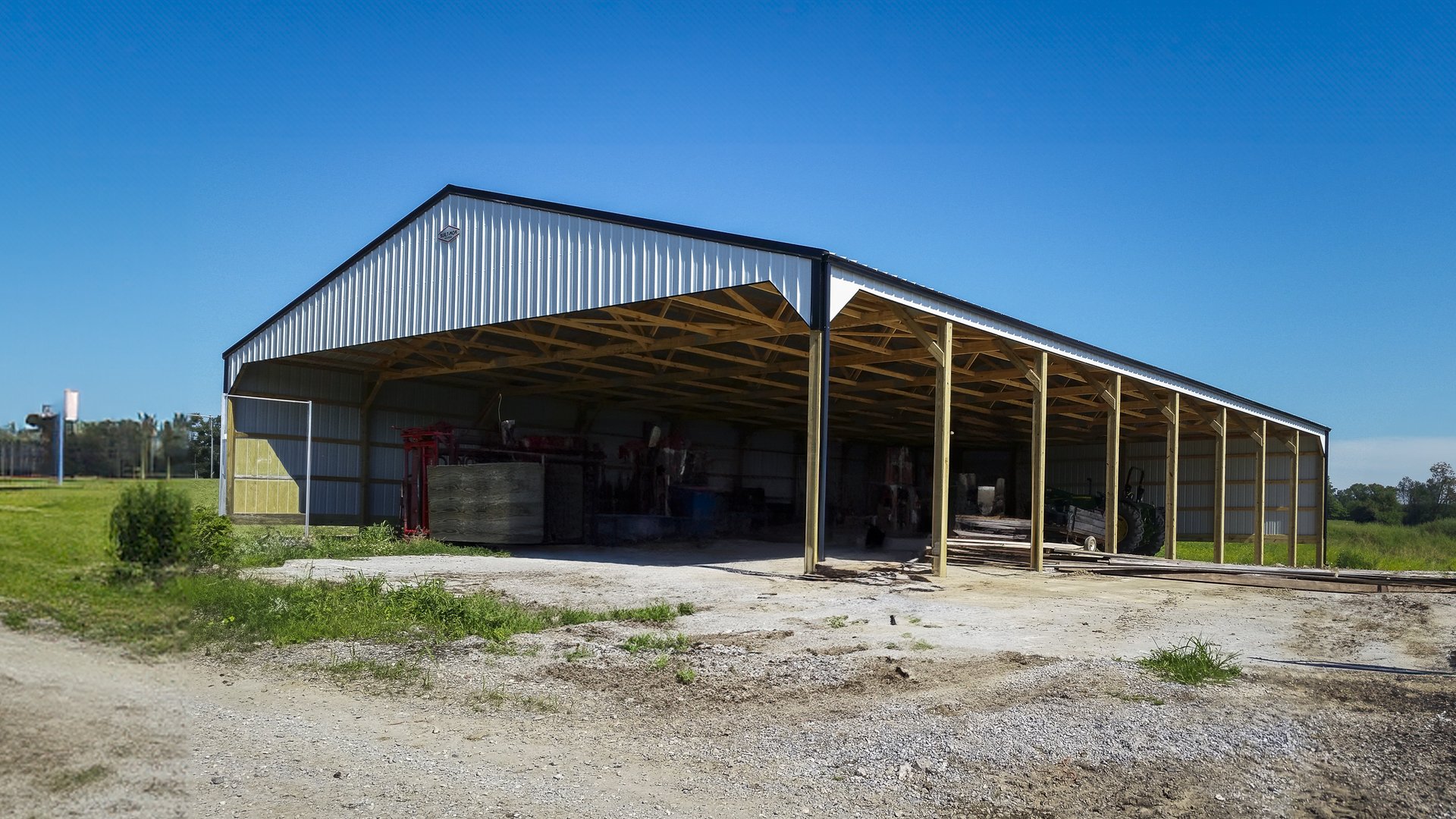 The image depicts a large post-frame construction building, likely a storage or agricultural facility, with an open side displaying a spacious interior. The structure features a metal roof and siding, supported by wooden posts and beams. The roof has a ga