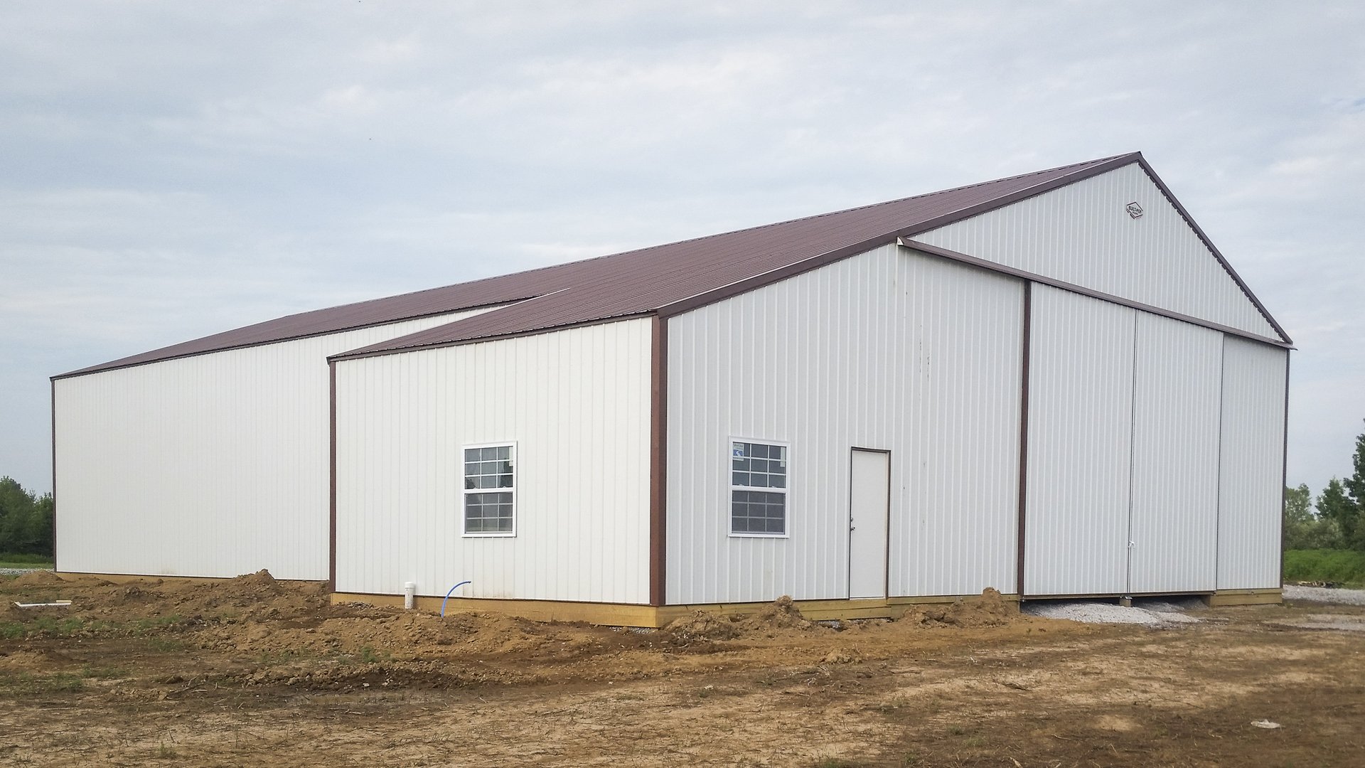 The image shows a post-frame construction building with a gable roof, featuring brown trim and white metal siding. The structure includes multiple sections, with the main section having large sliding doors. Two smaller windows and a single door are visibl