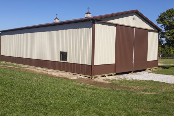 This image shows a post-frame building from Built-Mor Buildings, featuring a rectangular design with beige and brown vertical siding. The structure has a gabled roof with two cupolas. A large sliding door is located at the front, and a small window is pre