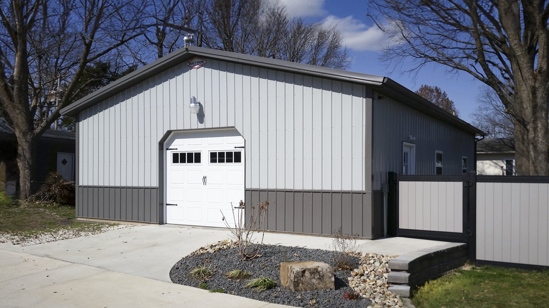 The image features a post-frame construction building, likely used as a garage or storage facility. It has a rectangular shape with a gable roof, finished in two-tone metal siding—light gray on the upper section and dark gray on the lower. A prominent whi