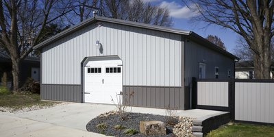 The image features a post-frame construction building, likely used as a garage or storage facility. It has a rectangular shape with a gable roof, finished in two-tone metal siding—light gray on the upper section and dark gray on the lower. A prominent whi