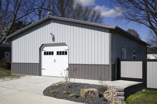 The image features a post-frame construction building, likely used as a garage or storage facility. It has a rectangular shape with a gable roof, finished in two-tone metal siding—light gray on the upper section and dark gray on the lower. A prominent whi