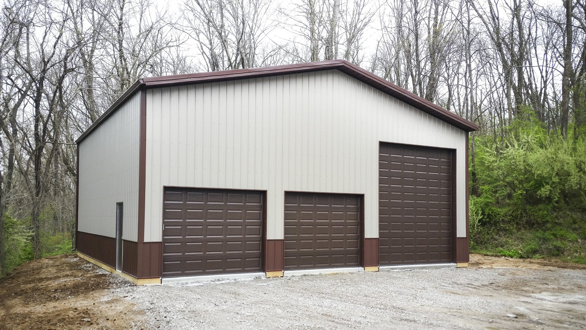 The image shows a post-frame construction building by Built-Mor Buildings, featuring a metal exterior with a light grey color and contrasting dark brown trim. It has three large overhead garage doors in the front, suggesting a purpose for vehicle storage