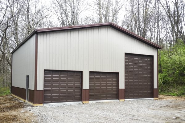 The image shows a post-frame construction building by Built-Mor Buildings, featuring a metal exterior with a light grey color and contrasting dark brown trim. It has three large overhead garage doors in the front, suggesting a purpose for vehicle storage