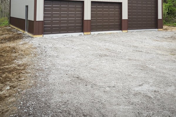 A post-frame building with a beige and brown exterior, featuring three large garage doors. The structure, resembling a storage or workshop facility, is situated on a gravel lot surrounded by trees. Its rectangular design and prominent overhead doors sugge