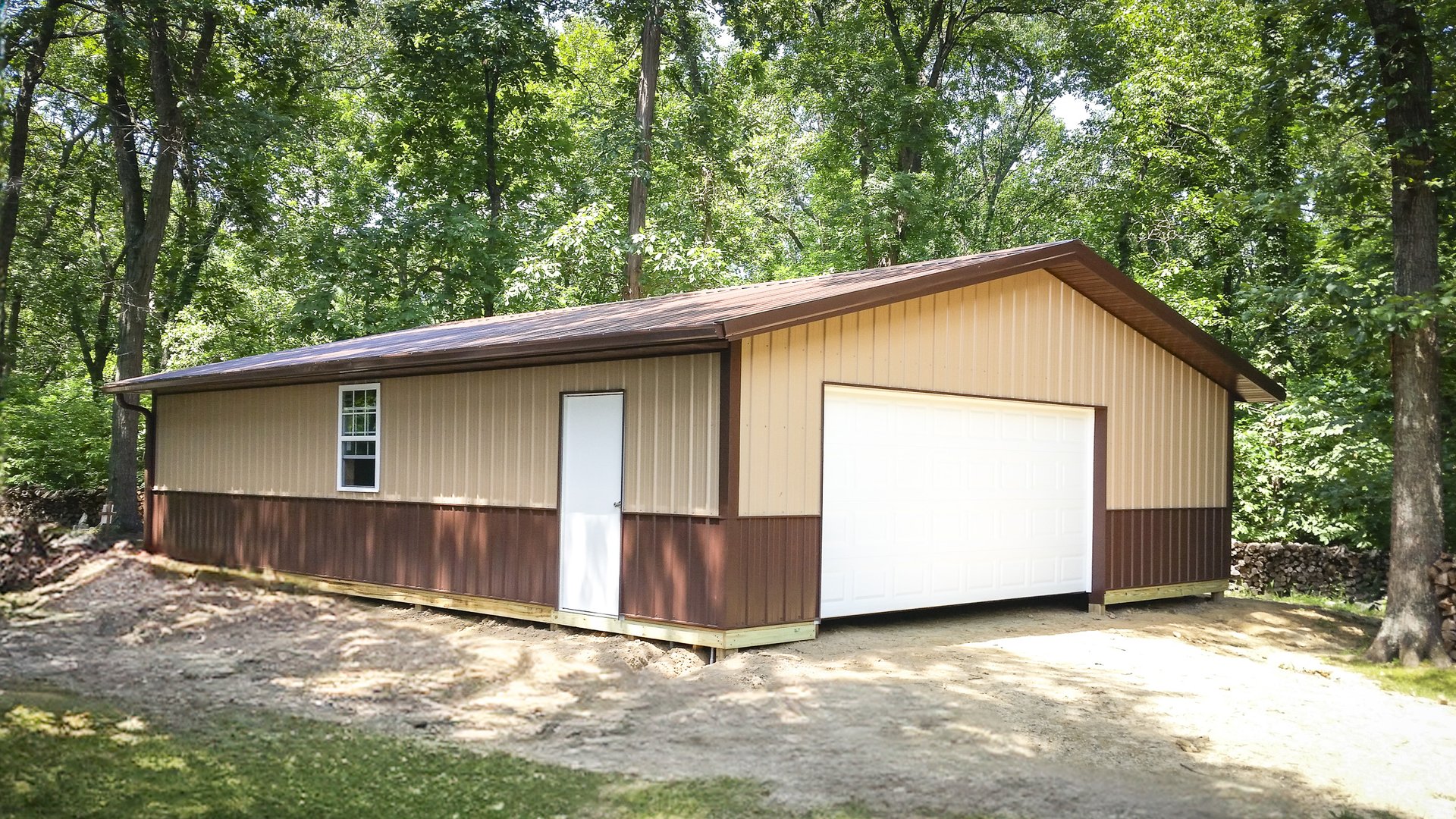 A post-frame building set in a wooded area, featuring a beige and brown exterior with a gabled roof. It has a large white garage door on the front and a single white pedestrian door on the side, next to a window. The structure appears to be used for stora