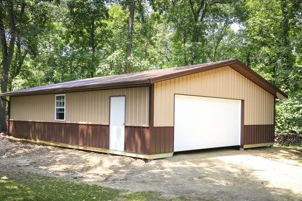 A post-frame building set in a wooded area, featuring a beige and brown exterior with a gabled roof. It has a large white garage door on the front and a single white pedestrian door on the side, next to a window. The structure appears to be used for stora