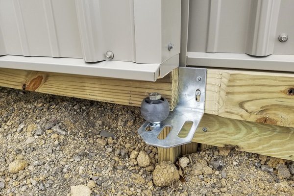 Close-up of a post-frame construction showing a corner of a building from Built-Mor Buildings. The image highlights key structural elements, including a metal siding panel attached to a treated wooden baseboard. A metal bracket with a rubber stopper suppo