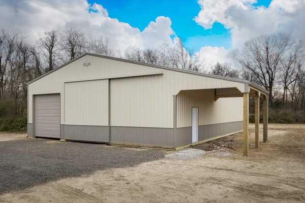 A light-colored post-frame building is shown with a gable roof and vertical siding. It features a large overhead garage door on the front, ideal for vehicle storage or a workshop. A smaller pedestrian door is present on the side, under a canopy supported