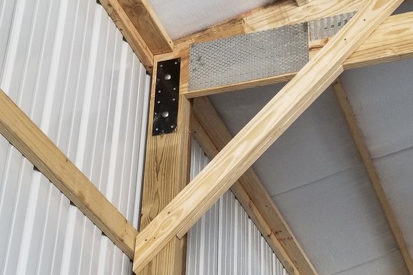 The image shows a detailed view of a post-frame construction corner from Built-Mor Buildings. It features wooden beams with metal connectors and braces for structural support. The interior wall is lined with vertical white metal panels, and insulation is