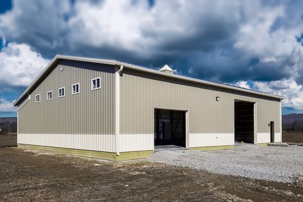 The image shows a large post-frame construction by Built-Mor Buildings, situated on a flat, open field. The building features a beige exterior with light-colored accents at the bottom. It has a gabled roof with a small cupola on top, adding a touch of arc