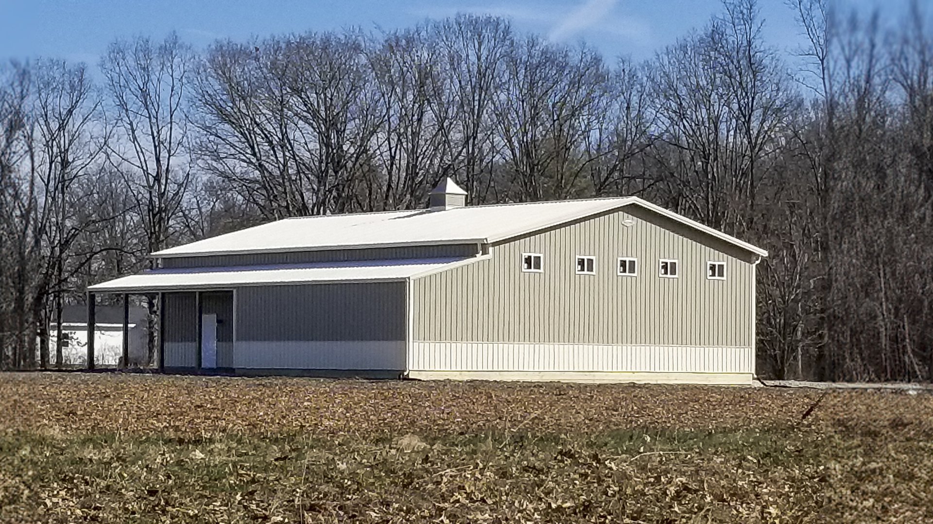 The image features a post-frame construction building from Built-Mor Buildings, designed for agricultural purposes. It has a light gray exterior with white trim and a gabled roof. There are small square windows along the side, and a large sliding door at