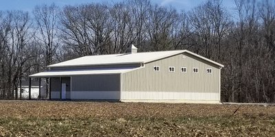 The image features a post-frame construction building from Built-Mor Buildings, designed for agricultural purposes. It has a light gray exterior with white trim and a gabled roof. There are small square windows along the side, and a large sliding door at