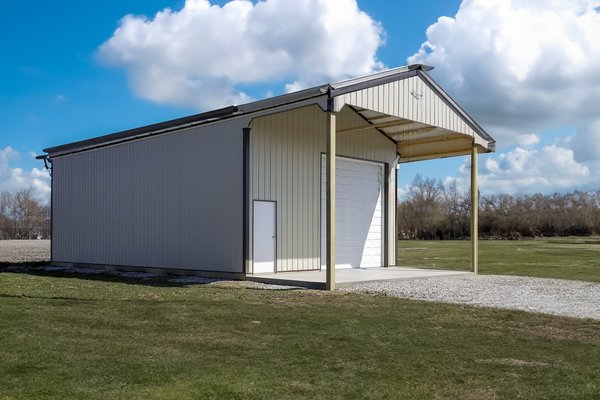 This image shows a post-frame building with beige metal siding and a gabled roof, featuring a large roll-up garage door at the front. The structure includes a small adjacent side door and an extended covered porch supported by two vertical posts. The buil