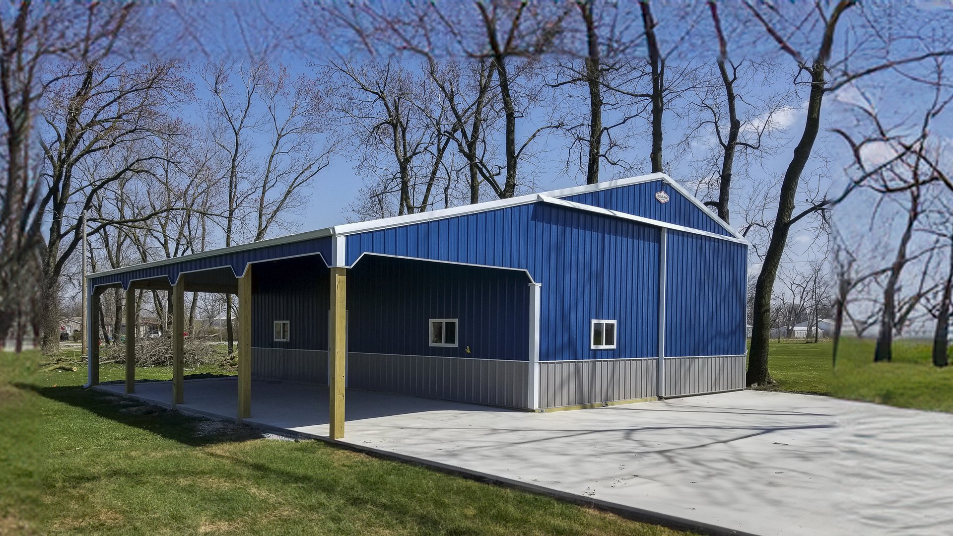 A blue post-frame building by Built-Mor Buildings features a gabled roof and a combination of metal siding in blue and light gray. The structure includes a covered open area supported by wooden posts, likely used as a carport or patio. The building is sit