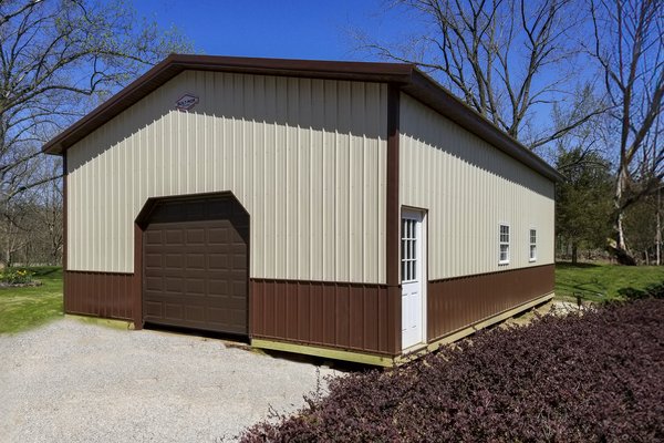 The image features a post-frame construction building by Built-Mor Buildings. It is a rectangular structure with a gable roof, designed for storage or workshop use. The exterior is clad in vertical metal siding, with the upper section in cream and the low