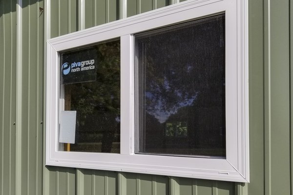 Close-up image of a post-frame building featuring a green metal siding with vertical ridges. A white-framed rectangular window is centrally positioned, with one section containing a mesh screen. Inside the window, signage for “pivagroup north america” is