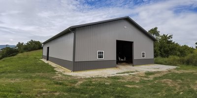 The image shows a post-frame construction building by Built-Mor Buildings, featuring a simple gable roof and metal siding. The exterior is divided into two tones, with darker panels at the bottom and lighter panels above. The structure includes two small