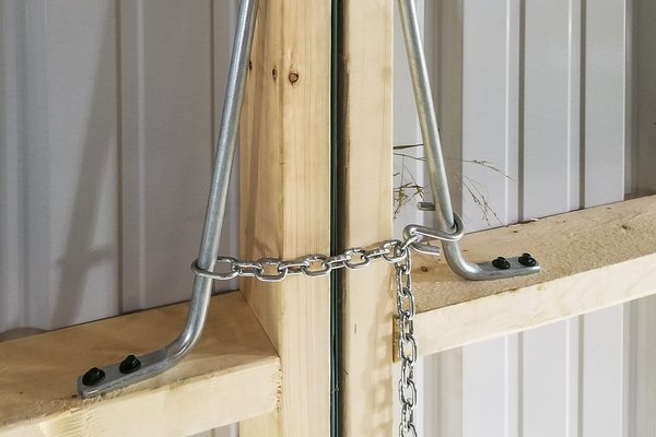 The image shows a close-up of a post-frame construction element from Built-Mor Buildings, highlighting a wooden post and horizontal beam joint. Metal brackets with bolts secure the beams, and a chain is looped for additional reinforcement. The background