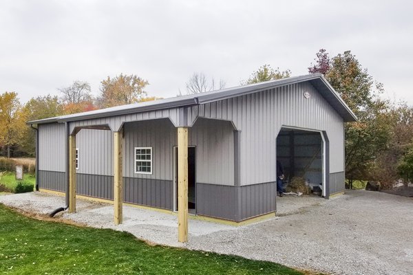 The image depicts a post-frame construction building by Built-Mor Buildings, primarily made of steel with a gabled roof. The structure features a large open bay on one side, indicating its use as a storage or workspace, likely a garage or workshop. It has