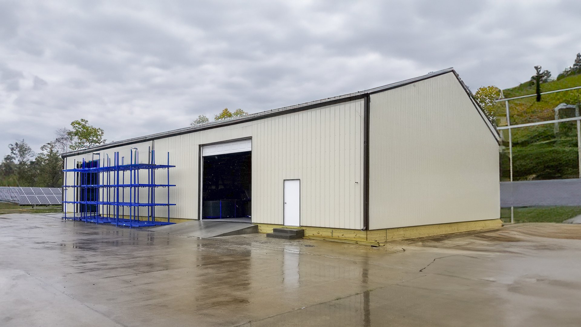 The image depicts a post-frame construction building designed for industrial or storage purposes. The structure features a peaked roof and light-colored metal siding. It includes a large, open sliding door for vehicle or equipment access and a smaller per