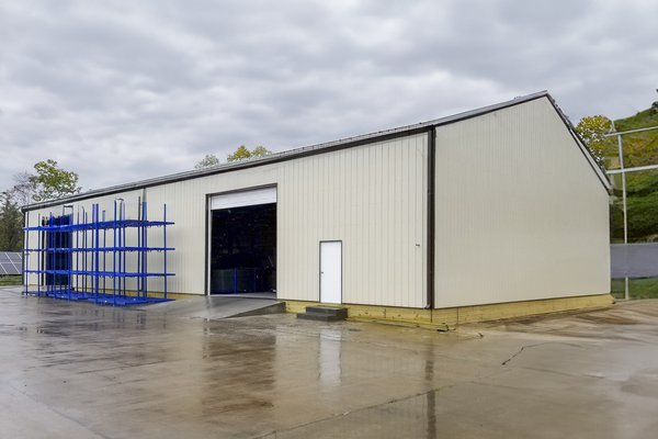 The image depicts a post-frame construction building designed for industrial or storage purposes. The structure features a peaked roof and light-colored metal siding. It includes a large, open sliding door for vehicle or equipment access and a smaller per