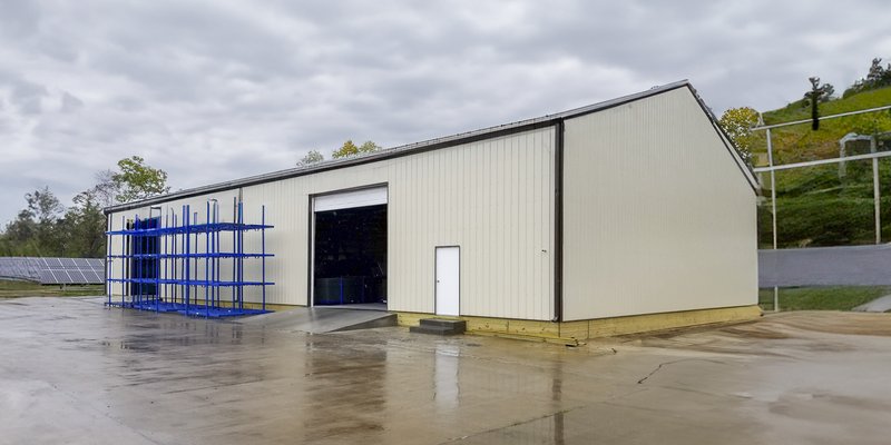 The image depicts a post-frame construction building designed for industrial or storage purposes. The structure features a peaked roof and light-colored metal siding. It includes a large, open sliding door for vehicle or equipment access and a smaller per