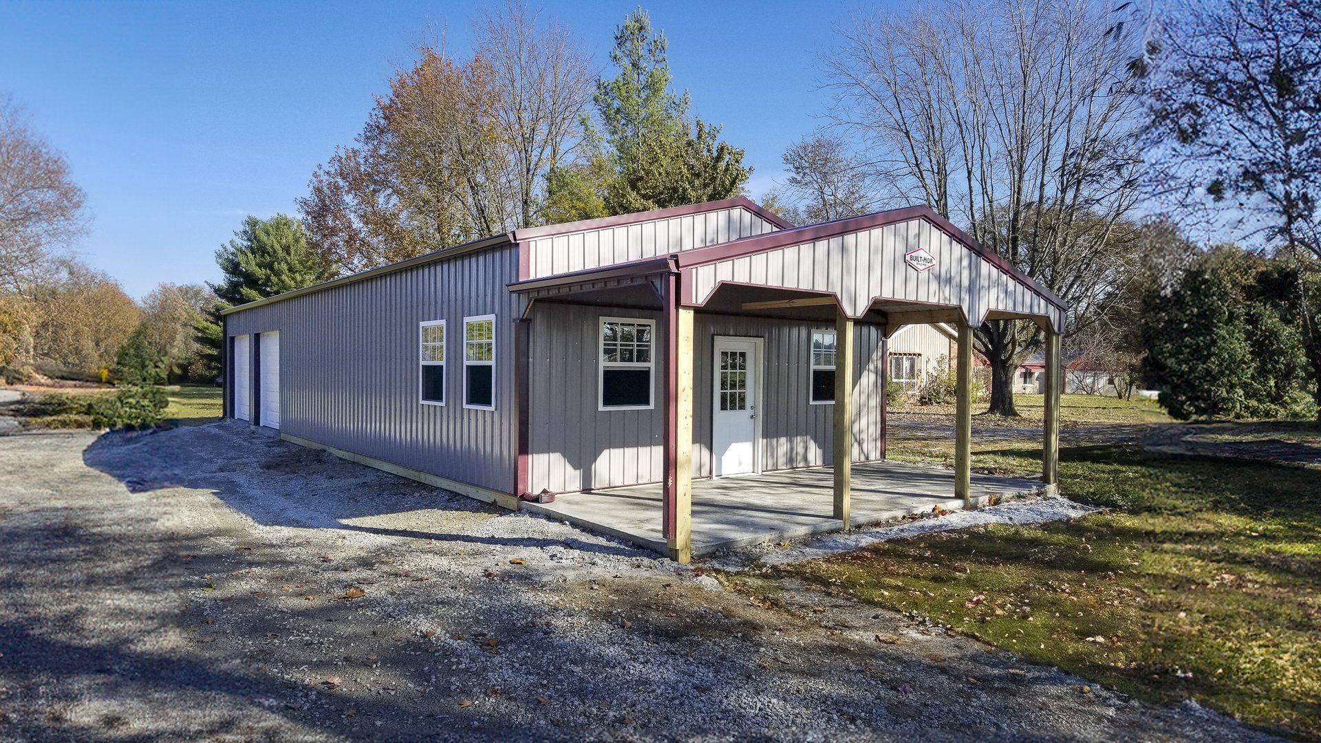 The image shows a post-frame building from Built-Mor Buildings, featuring metal siding and roofing with light gray and maroon trim. It has a gable-style roof and an extended entrance porch supported by wooden posts. The structure includes multiple windows