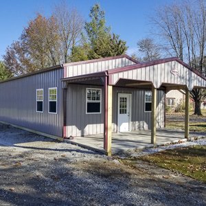 The image shows a post-frame building from Built-Mor Buildings, featuring metal siding and roofing with light gray and maroon trim. It has a gable-style roof and an extended entrance porch supported by wooden posts. The structure includes multiple windows