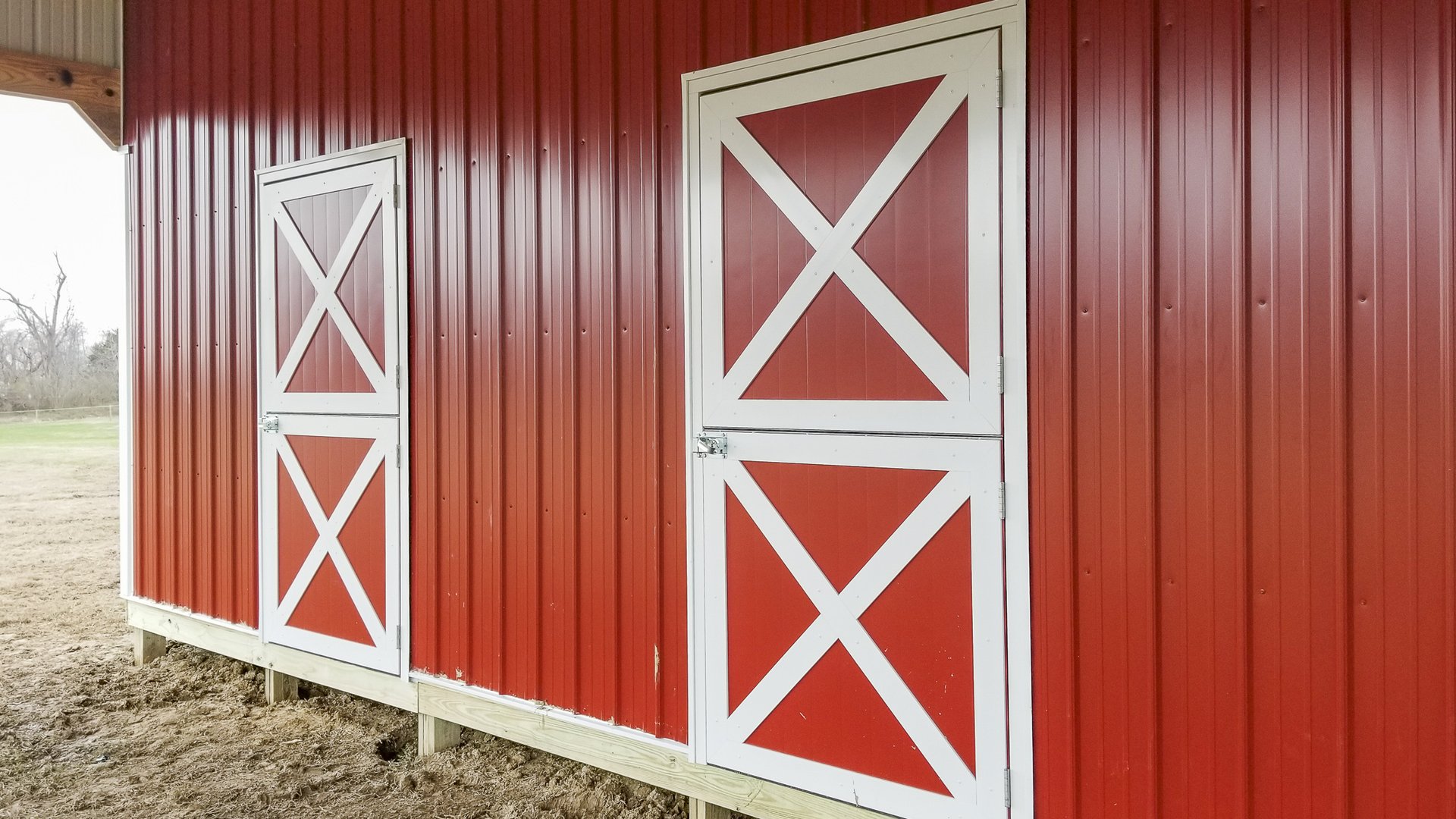 The image shows a post-frame building by Built-Mor Buildings, featuring red vertical metal siding with white diagonal cross-braced doors, typical of barn-style design. The exterior wall includes two doors of different sizes, both secured with metal latche