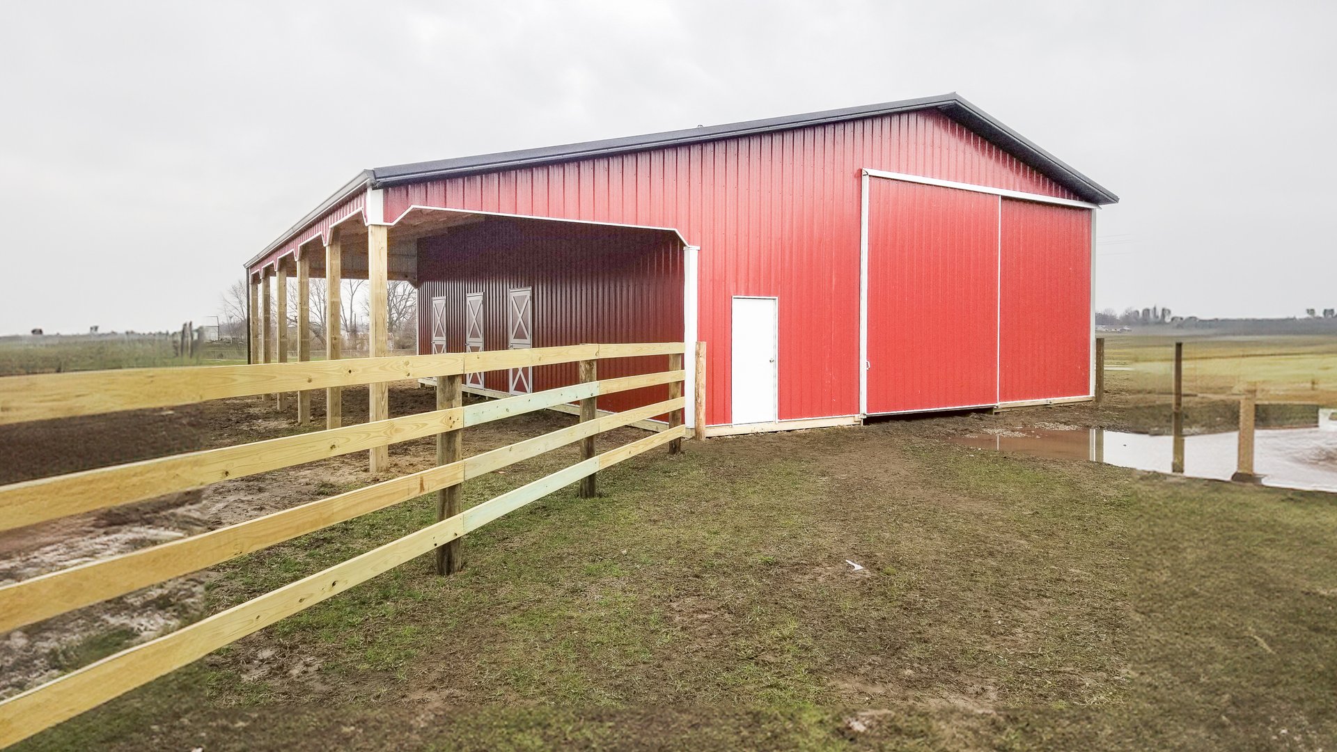 The image shows a post-frame construction building by Built-Mor Buildings, featuring a red exterior with a white door and large sliding barn-style doors. The building has a gable roof and an open covered area on one side, supported by wooden posts. It is