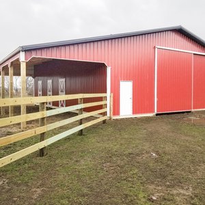 The image shows a post-frame construction building by Built-Mor Buildings, featuring a red exterior with a white door and large sliding barn-style doors. The building has a gable roof and an open covered area on one side, supported by wooden posts. It is