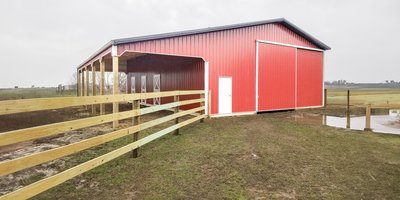 The image shows a post-frame construction building by Built-Mor Buildings, featuring a red exterior with a white door and large sliding barn-style doors. The building has a gable roof and an open covered area on one side, supported by wooden posts. It is