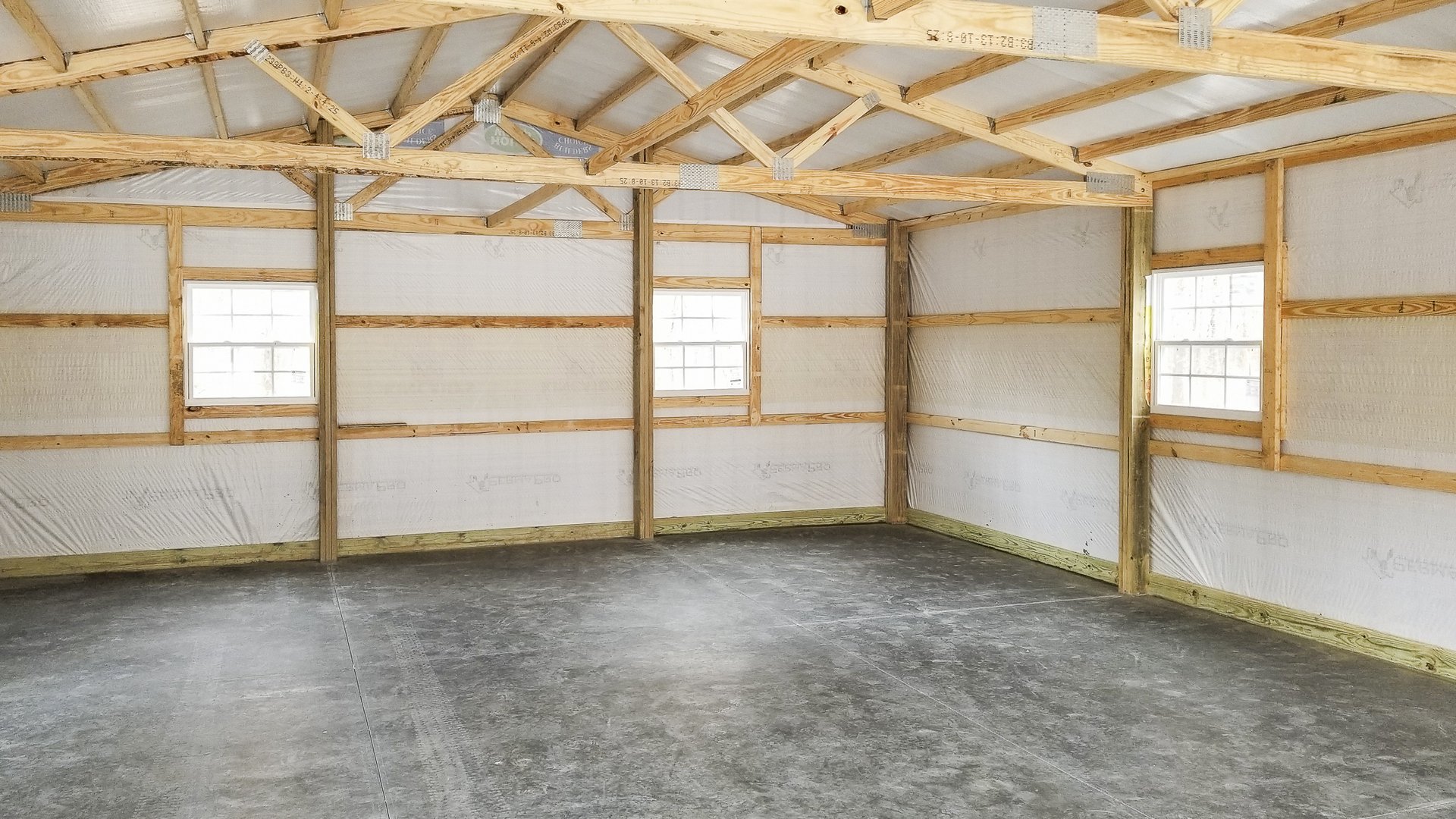 Interior of a post-frame building by Built-Mor Buildings, featuring exposed wooden trusses and beams supporting a pitched roof. The walls are lined with insulation panels and framed with horizontal wood members. The floor is concrete, suggesting use for s