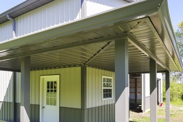 The post-frame construction image from Built-Mor Buildings shows a metal building with a prominent covered porch area. The structure features light gray siding with darker gray trim and roofing, supported by sturdy metal posts. A white entry door with a w