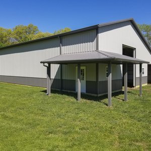 The image shows a large post-frame construction building by Built-Mor Buildings, featuring a dual-tone exterior with light gray upper walls and darker gray lower sections. A prominent feature is the covered porch with a sloped roof supported by wooden pos