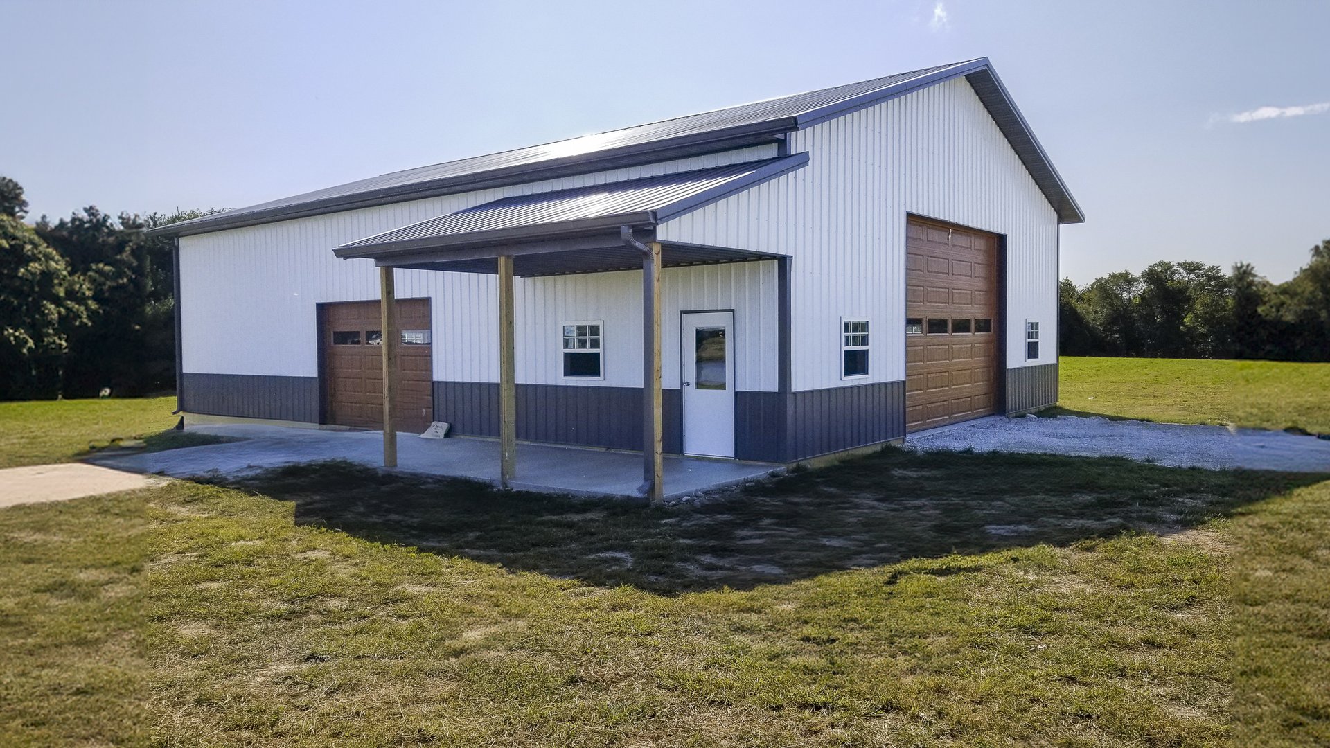 The image depicts a post-frame building from Built-Mor Buildings, featuring a gable roof and metal siding in white and gray. The structure has three large overhead doors, suggesting use as a garage or storage facility. A small covered entryway, supported