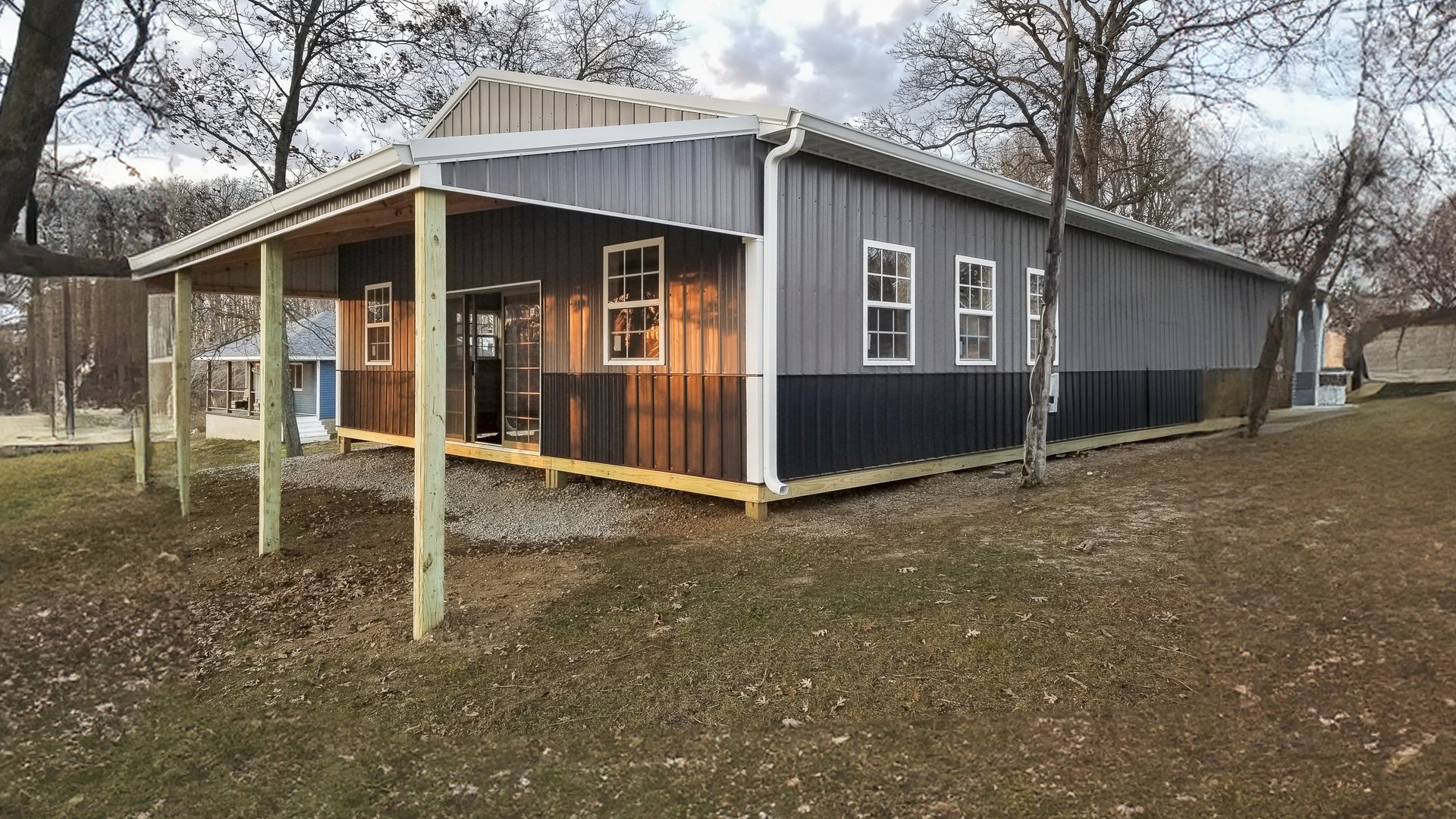 The image shows a post-frame construction building from Built-Mor Buildings. It features a gable roof and an extended covered porch area supported by wooden posts. The exterior walls have a two-tone design with vertical siding, light gray on the upper hal