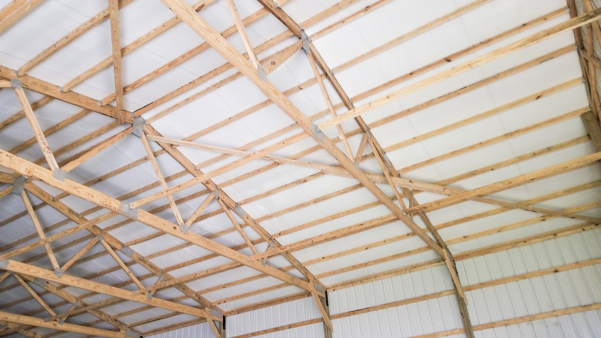 This image shows the interior of a post-frame construction building by Built-Mor Buildings. The structure features wooden trusses with metal connectors supporting a white ceiling. The walls are lined with vertical metal panels. The trusses are arranged in