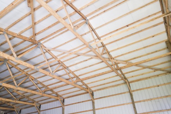 This image shows the interior of a post-frame construction building by Built-Mor Buildings. The structure features wooden trusses with metal connectors supporting a white ceiling. The walls are lined with vertical metal panels. The trusses are arranged in