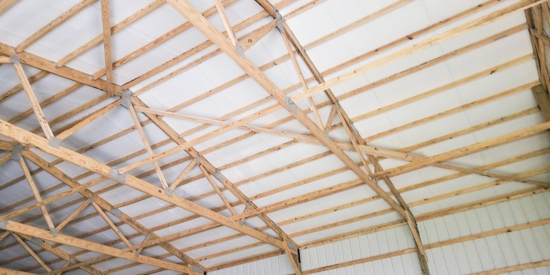 This image shows the interior of a post-frame construction building by Built-Mor Buildings. The structure features wooden trusses with metal connectors supporting a white ceiling. The walls are lined with vertical metal panels. The trusses are arranged in