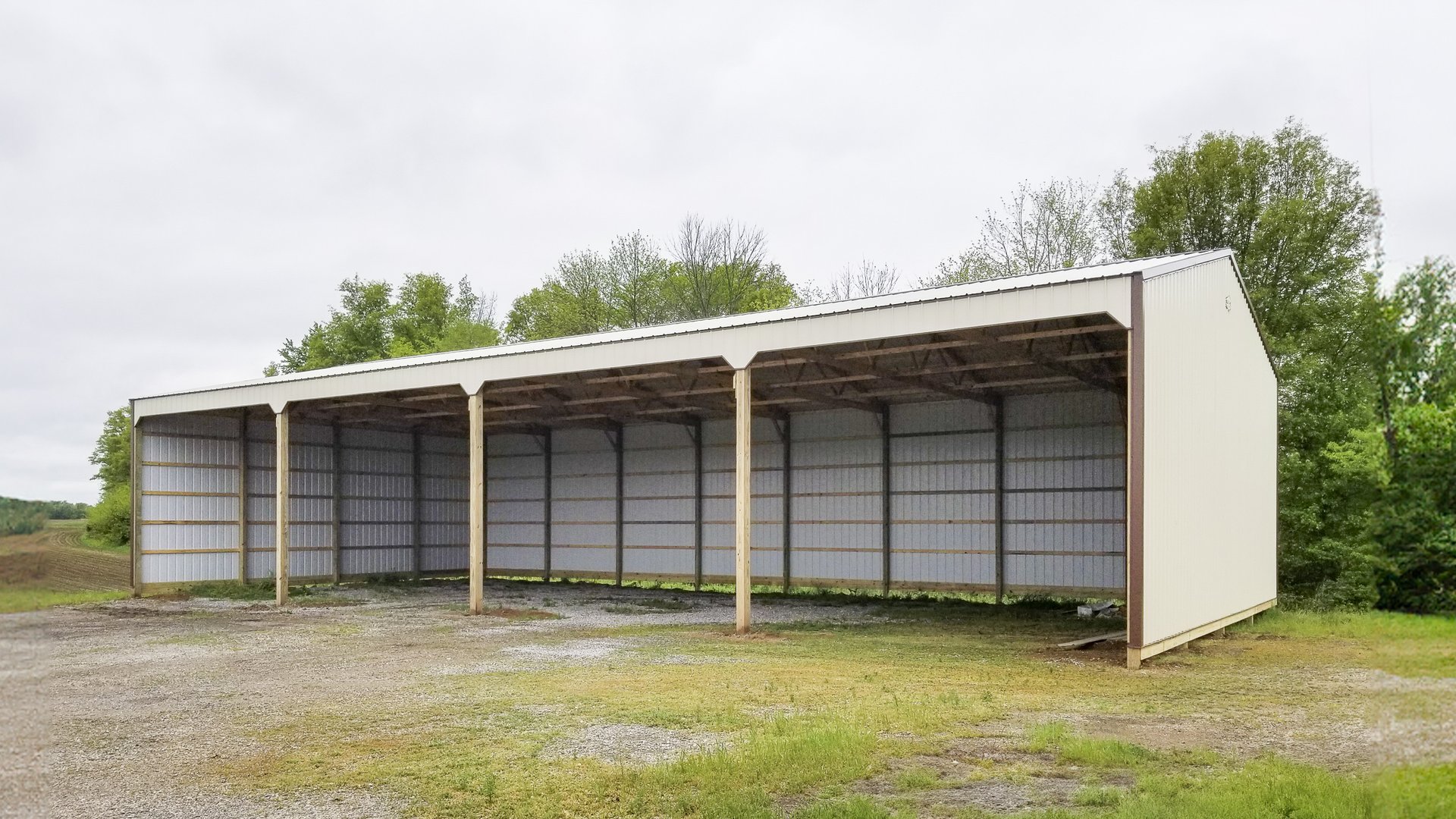 The image depicts a post-frame construction by Built-Mor Buildings, featuring a large open-sided structure likely used for equipment or vehicle storage. The building has a metal roof supported by wooden posts, with three open bays, each divided by vertica