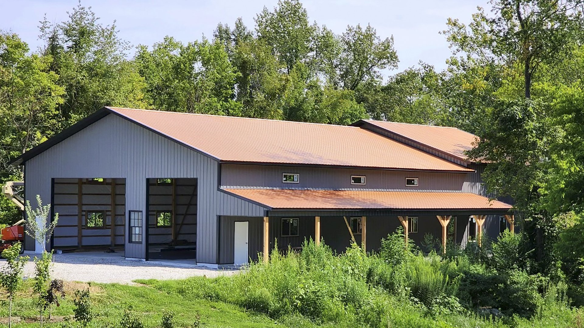 The image shows a post-frame building by Built-Mor Buildings, featuring a large, rectangular structure with a pitched metal roof. The building has two distinct sections: a taller main area with large open bays, likely for storage or workspace, and a lower