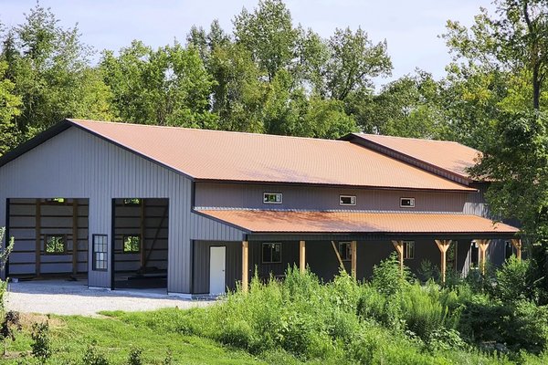 The image shows a post-frame building by Built-Mor Buildings, featuring a large, rectangular structure with a pitched metal roof. The building has two distinct sections: a taller main area with large open bays, likely for storage or workspace, and a lower