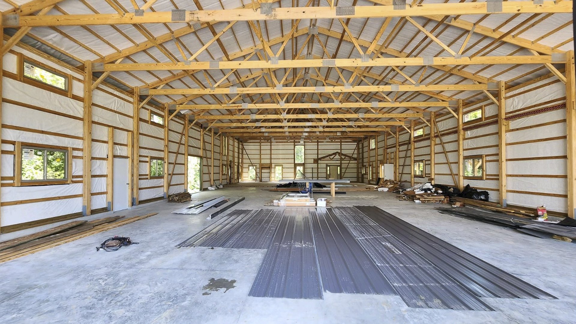 Interior view of a post-frame construction building by Built-Mor Buildings, featuring a spacious, open layout with exposed wooden trusses supporting the roof. The structure has insulated walls and large, evenly spaced windows allowing natural light. Build